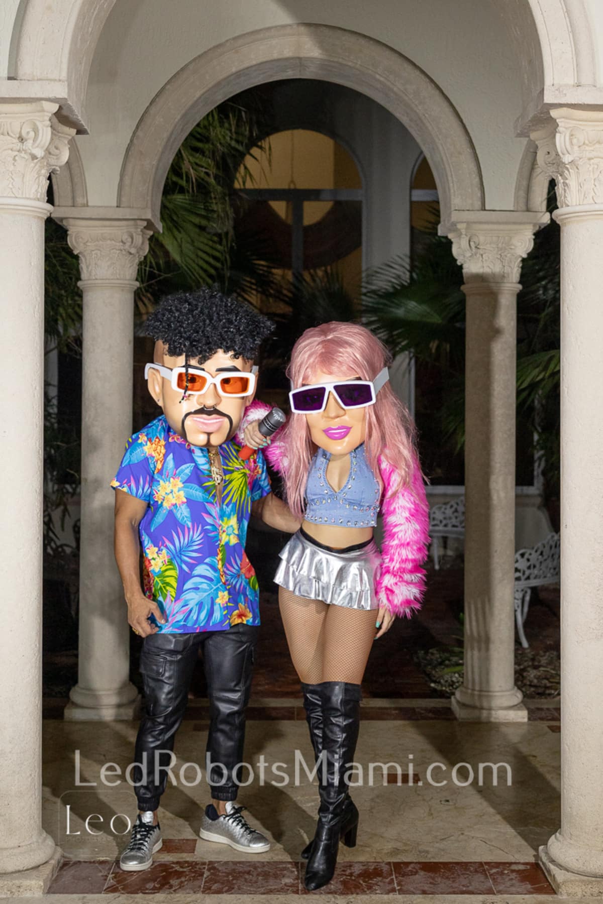 Hora Loca Service in Miami Two People Wear Large Cartoonish Masks and Colorful Outfits Standing Under an Archway One Wears a Floral Shirt and Leather Pants the Other a Pink Faux Fur Jacket and Metallic Skirt the Background Includes Greenery and White Columns