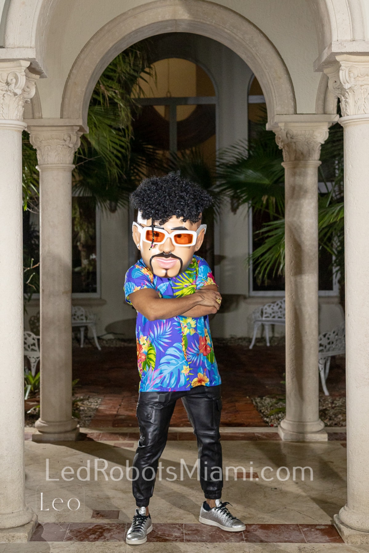 Hora Loca Service in Miami Person Wearing a Colorful Tropical themed Outfit and Large Cartoonish Headpiece Resembling a Face with Glasses and Curly Hair Stands with Arms Crossed Under a Stone Archway with Greenery in the Background