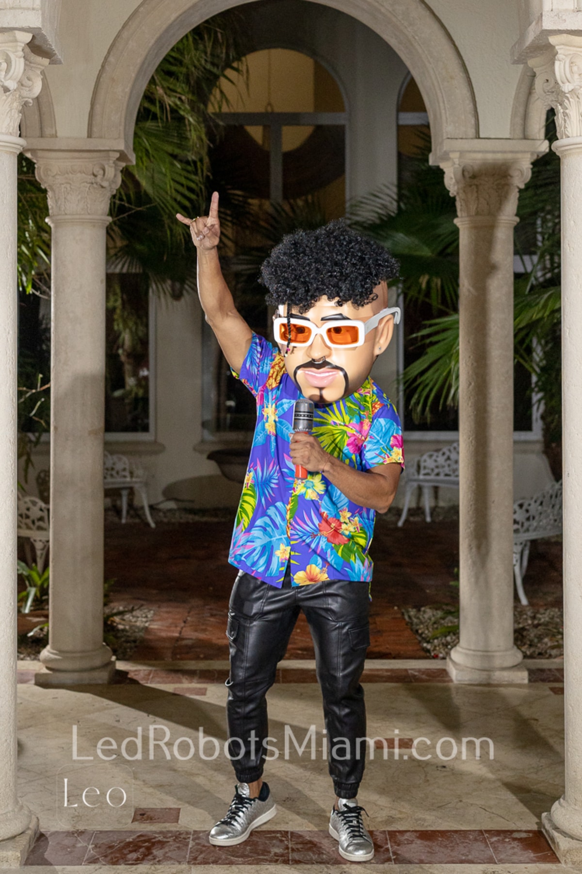 Hora Loca Service in Miami a Person Wearing a Tropical Shirt and Leather Pants Holding a Microphone Stands Confidently Under an Archway the Person is Wearing a Large Cartoon Style Mask with Curly Hair and Sunglasses the Setting Includes Lush Greenery